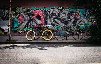 Two bicycles parked against a vibrant graffiti wall on a sunny day, adding urban flair. HD wallpaper
