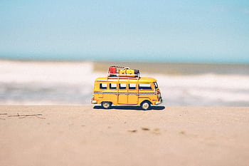 A vibrant yellow toy van sits on a sandy beach with an ocean backdrop, symbolizing fun travel adventures. HD wallpaper