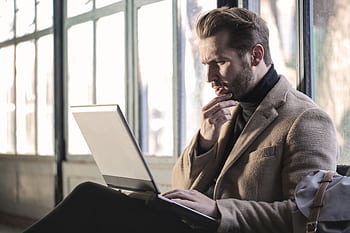 Man looking contemplative while working on a laptop in a well-lit indoor space. HD wallpaper