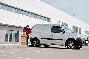 A delivery driver wearing a face mask unloads packages from a van outside a modern building. HD wallpaper