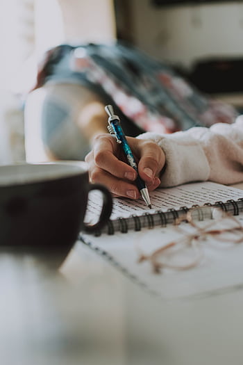 A person writes in a notebook while a cup of coffee sits nearby, indoors. HD phone wallpaper