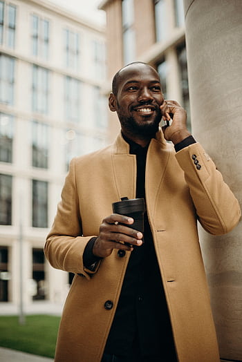 Smiling businessman in a trench coat holding a coffee cup while talking on the outdoors. HD phone wallpaper