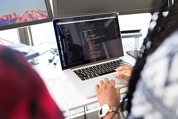 A woman coding on a laptop in a modern office environment with multiple monitors. HD wallpaper