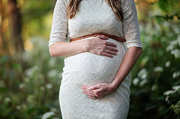 A pregnant woman in a lace dress gently cradling her belly in an outdoor setting. HD wallpaper