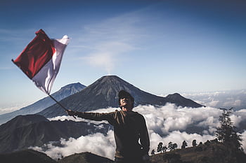 A hiker waves the Indonesian flag atop a scenic mountain, surrounded by clouds. HD wallpaper