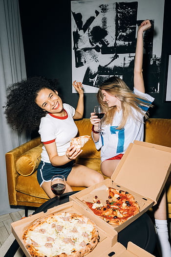 Two friends enjoying a fun pizza party indoors, sharing laughter and red wine. HD phone wallpaper