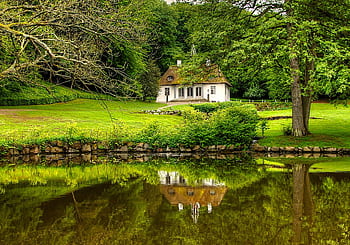 que cottage with a thatched roof reflecting on a serene lake surrounded by lush greenery. HD wallpaper