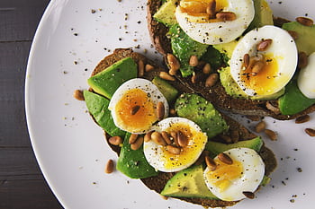 Delicious avocado toast topped with soft-boiled eggs and pine nuts on a white plate. HD wallpaper