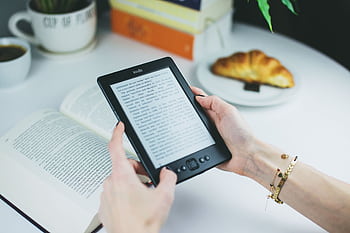 A woman holds a Kindle while reading with a book and croissant on a table. HD wallpaper