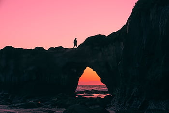 A person silhouetted on a coastal rock arch after sunset with a vibrant sky. HD wallpaper