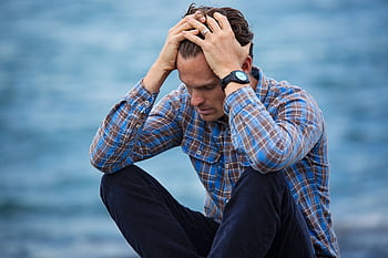 A man in a plaid shirt sits by the water looking distressed, symbolizing stress. HD wallpaper