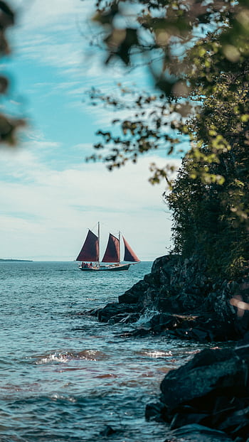 A serene seascape featuring a sailboat with red sails navigating along a rocky coastline. HD phone wallpaper