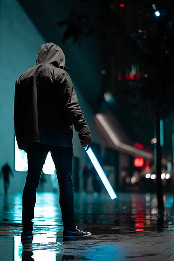 Silhouetted person in hoodie holding glowing light stick on rainy city street at night. HD phone wallpaper