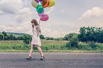A woman in a white dress walks on a road carrying colorful balloons against a scenic outdoor backdrop. HD wallpaper