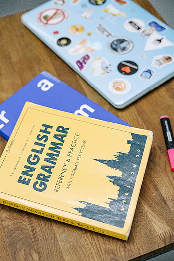 High-angle view of an English grammar book and a laptop with stickers on a wooden desk. HD phone wallpaper