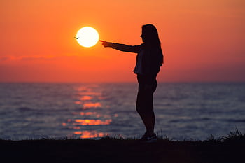 A woman silhouetted against a vibrant sunset at the beach, pointing at the sun with a bird in flight. HD wallpaper