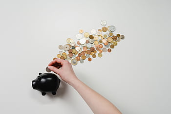 Person placing coin into a black piggy bank with scattered coins, symbolizing savings and finance. HD wallpaper