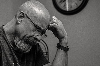 Black and white portrait of a thoughtful bald man indoors, capturing a moment of reflection with a wall clock in the background. HD wallpaper