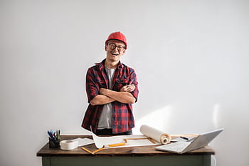 Smiling engineer in a red hardhat stands with arms crossed at a desk with blueprints and laptop. HD wallpaper