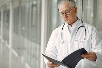 Elderly doctor with stethoscope attentively reading a medical report in a modern hospital setting. HD wallpaper
