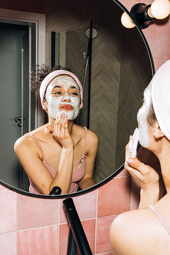 A young woman smiling while applying a facial mask during her home skincare routine. HD phone wallpaper