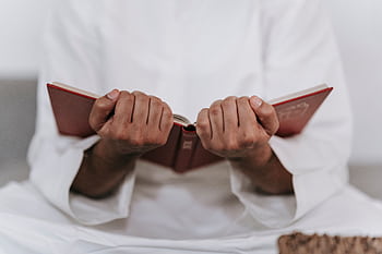 A man in traditional Islamic attire holds a Quran in a focused prayer session indoors. HD wallpaper