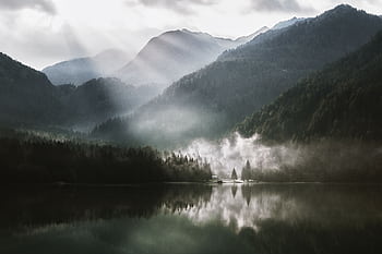 Breathtaking view of a foggy mountain landscape with sun rays piercing through, reflecting on a serene lake. HD wallpaper