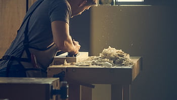 A craftsman skillfully shaping wood at a workshop bench with wood shavings visible. HD wallpaper