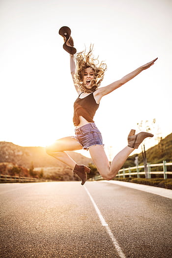 Happy woman jumping in the air on a sunny road with vibrant energy and joy. HD phone wallpaper