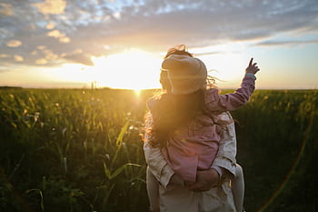 A mother and daughter embrace and point at the sunset in a grassy field. HD wallpaper