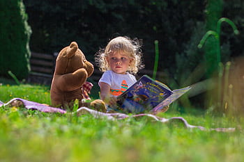 Young child reading a book outdoors with a teddy bear, enjoying a sunny day on the grass. HD wallpaper