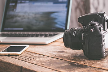 Close-up of a DSLR camera, laptop, and smart on a wooden table in a modern workspace. HD wallpaper