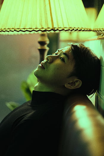 A contemplative Asian man leans on a sofa under warm lamp light, expressing solitude. HD phone wallpaper