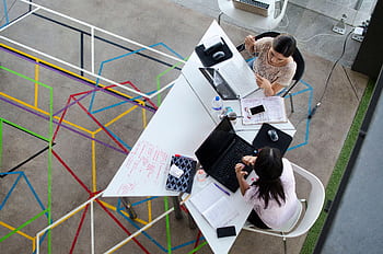 Overhead view of women working together in a modern office setting with colorful geometric floor design. HD wallpaper