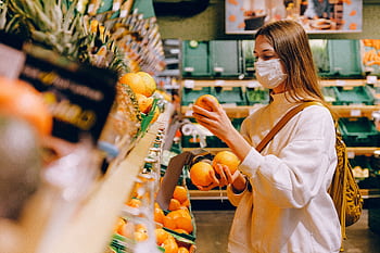 A woman wearing a face mask selects oranges in a grocery store, focusing on safety and prevention. HD wallpaper