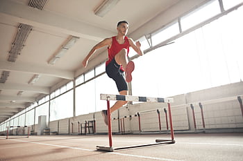 Dynamic shot of a male athlete in red sportswear jumping over a hurdle indoors. HD wallpaper