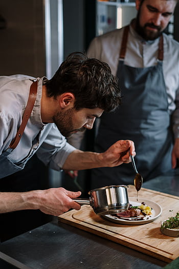 Two chefs in aprons skillfully plating a gourmet dish in an industrial kitchen setting. HD phone wallpaper