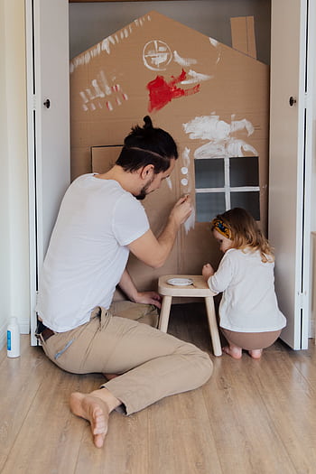 Father and daughter painting a cardboard house indoors, enjoying quality time together. HD phone wallpaper