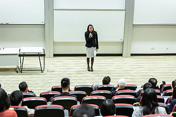 Female speaker presenting to an audience in a modern auditorium setting. HD wallpaper