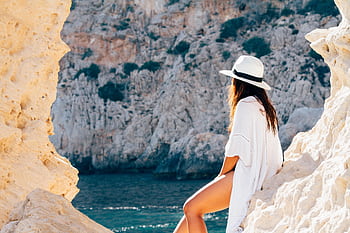 A woman in a hat sits on a rocky beach in summer, enjoying the sea view. HD wallpaper