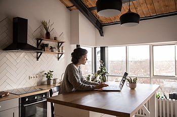 A young woman works on her laptop in a modern, bright kitchen setting with natural decor. HD wallpaper