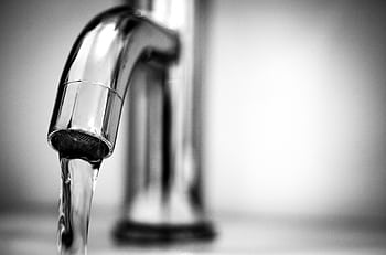 Black and white close-up of water flowing from a shiny stainless steel faucet indoors. HD wallpaper