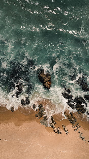 Stunning aerial shot capturing waves crashing on a sandy beach with exposed rocks, showcasing nature's raw beauty. HD phone wallpaper