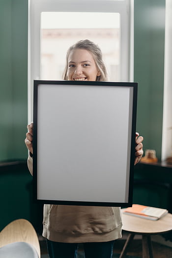 Smiling woman holding a blank frame in a cozy indoor setting, perfect for mockup design. HD phone wallpaper