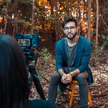 A young man being filmed outdoors in a forest, wearing casual attire and eyeglasses. HD phone wallpaper