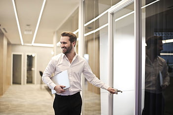 A smiling man holds a laptop while standing in a modern office hallway, exuding confidence. HD wallpaper
