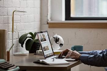 A tidy, modern workspace showing a person working on a laptop at a desk decorated with a globe, lamp, and plant. HD wallpaper