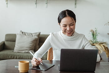 A cheerful woman uses a laptop and tablet for a video call, working remotely in a cozy living room. HD wallpaper