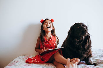 Joyful child in red dress laughing with a black dog indoors, capturing a heartwarming moment. HD wallpaper