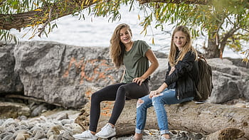 Two smiling teenage girls sitting on a log by the water, enjoying a casual day outdoors. HD wallpaper
